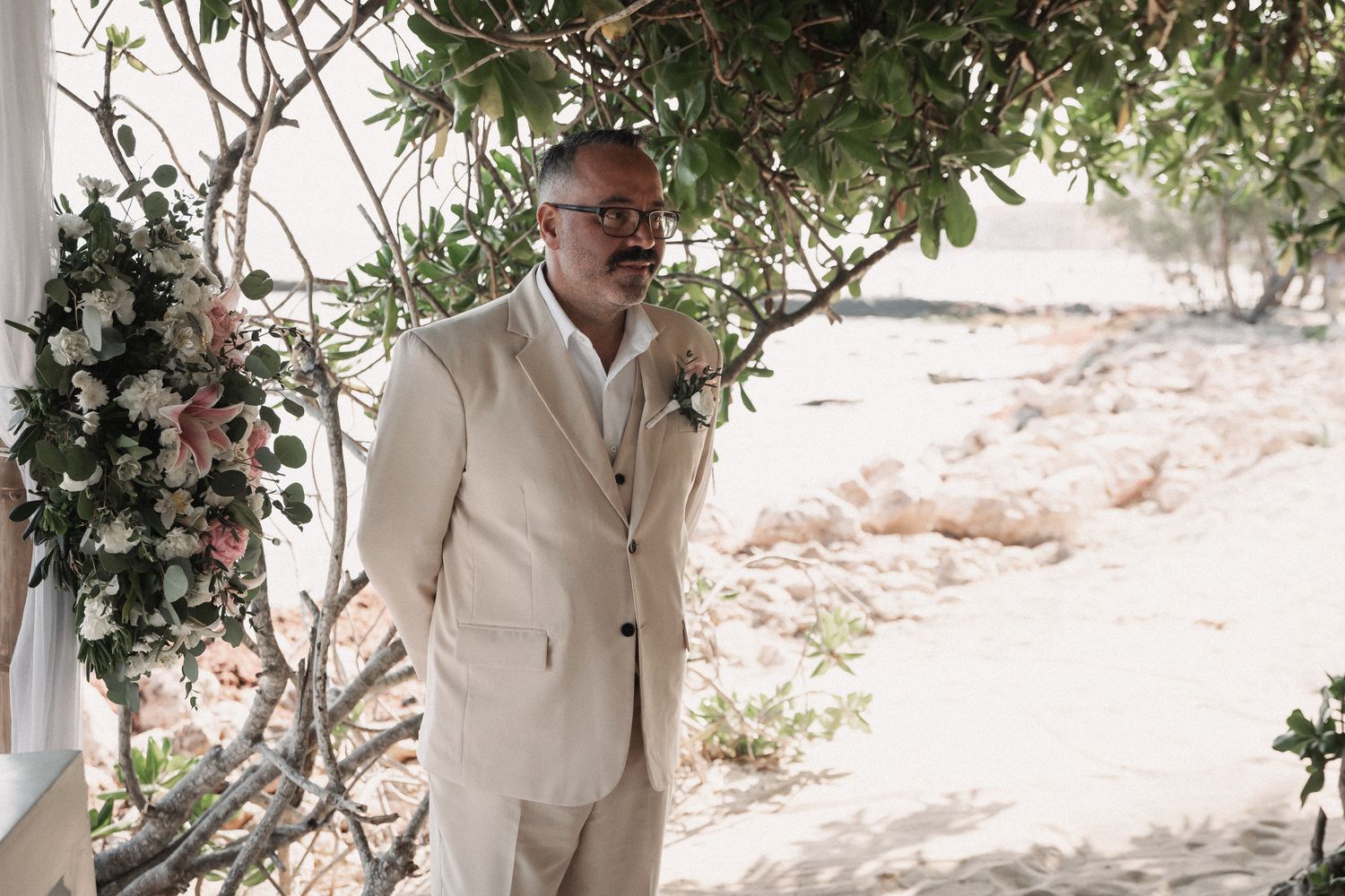 Groom in a beige linen suit standing beneath a floral arch on a sandy beach destination wedding venue.