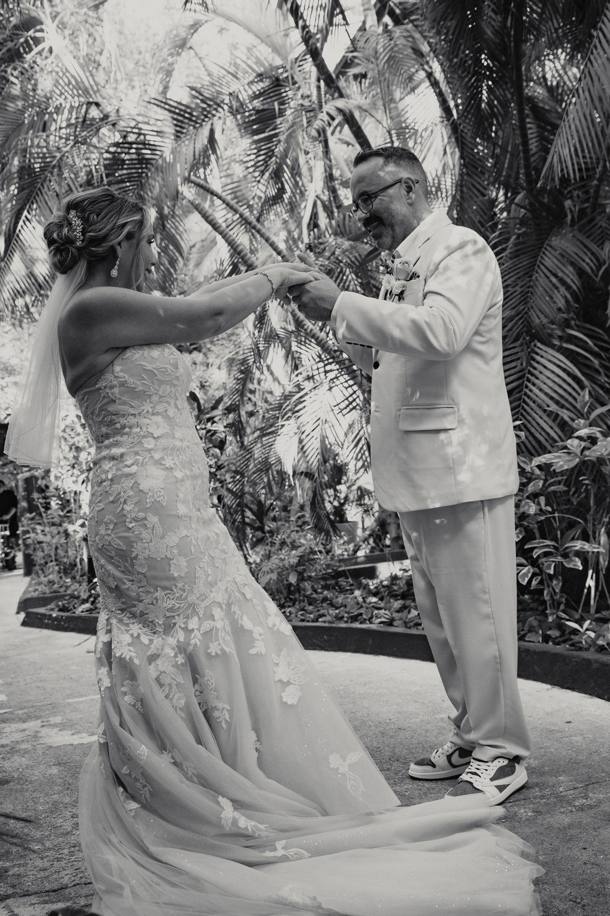 Black and white photo of bride and groom exchanging rings surrounded by tropical palms during ceremony.