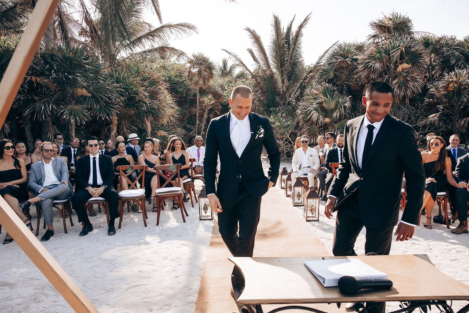 Wedding guests enjoy outdoor beach ceremony seating with palm trees and ocean views in the background.