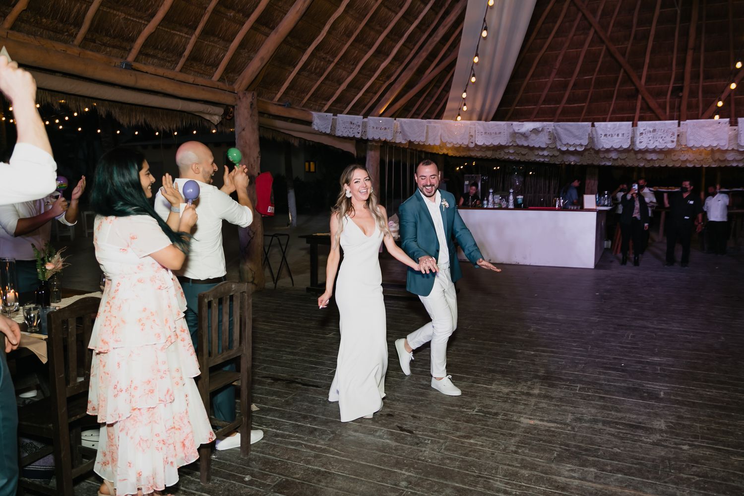 Bride and groom make joyful entrance into rustic outdoor wedding reception venue with string lights.
