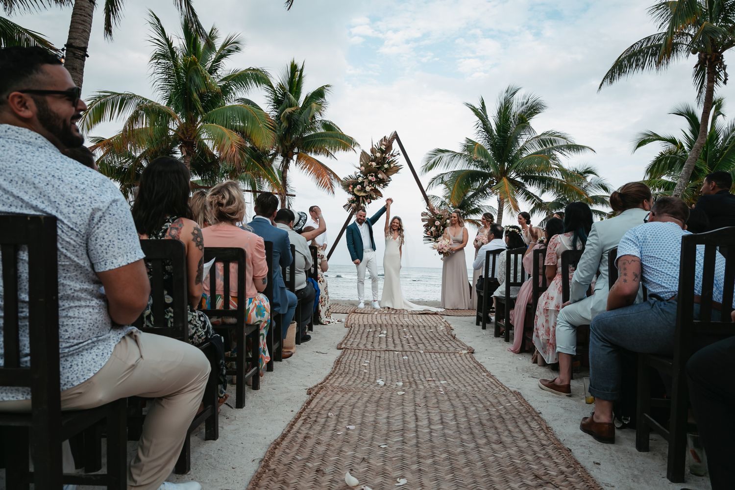 Romantic beach wedding ceremony with guests seated along a decorated aisle lined with palm trees.