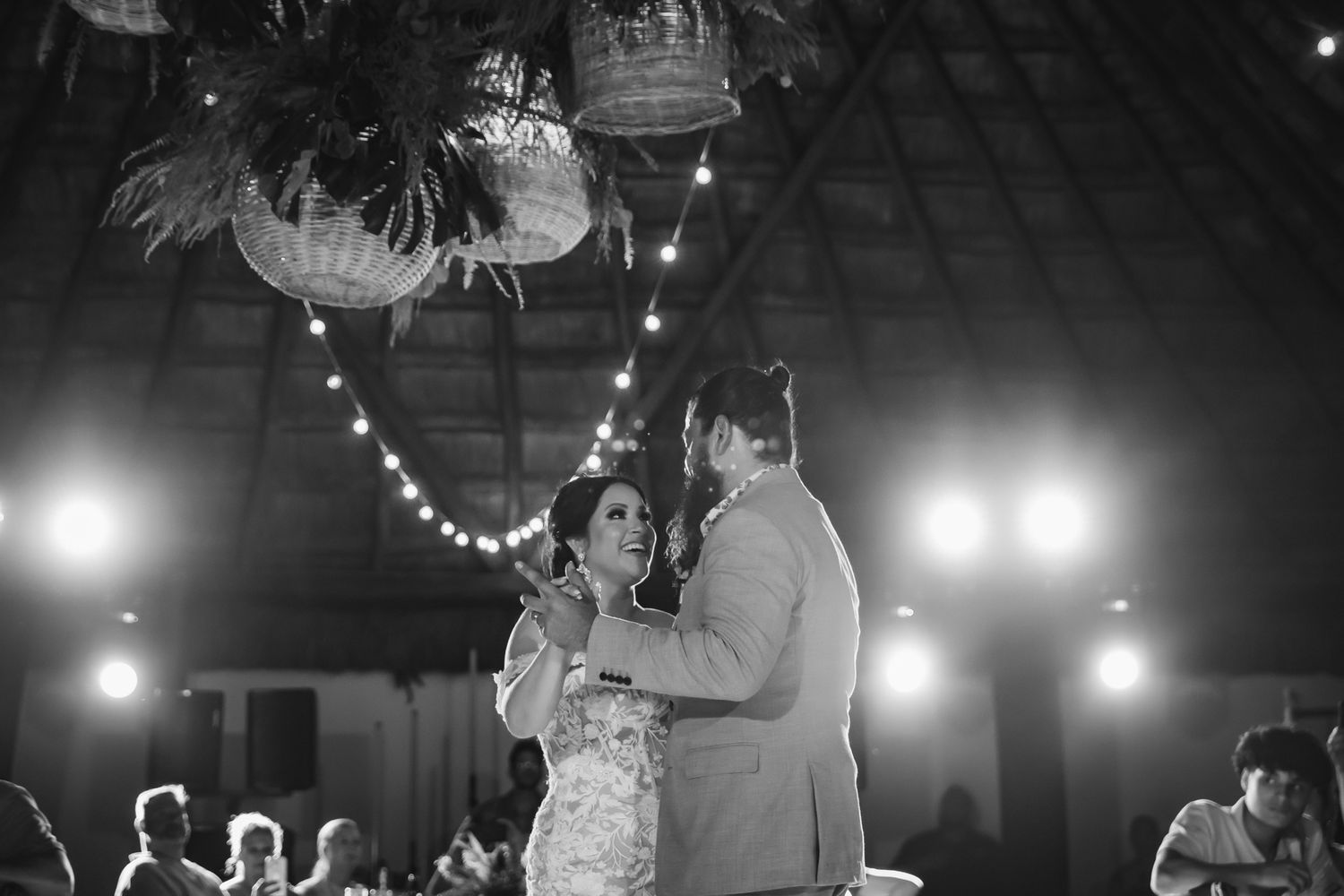 Black and white photo of a couple sharing their first dance at a wedding reception with string lights and floral decor.
