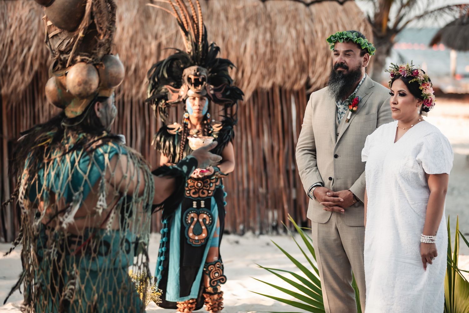 Couple stands at beach wedding altar with Mayan officiant in traditional costume and thatched hut backdrop.