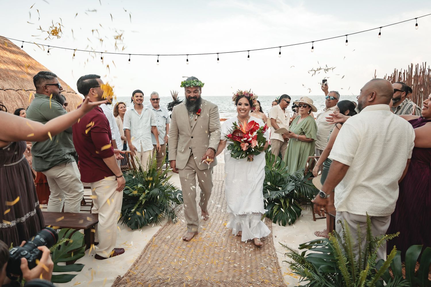Bride and groom walk down outdoor beach wedding aisle as guests celebrate with confetti and tropical floral decor.