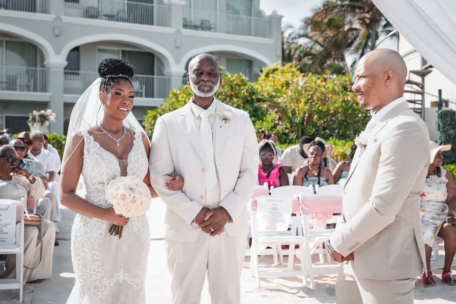 Bride in lace gown stands with groom and officiant at outdoor beach resort wedding ceremony.
