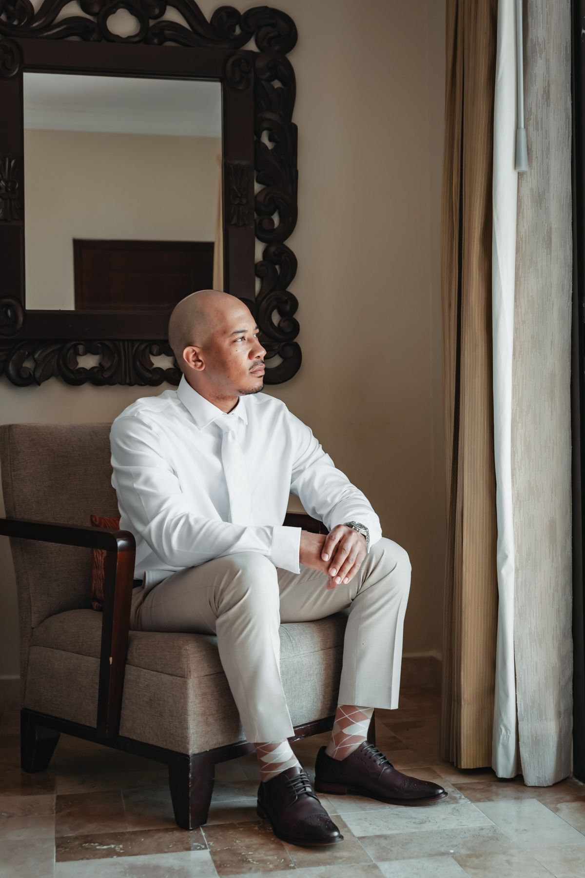 Bald man in white shirt and light pants sitting in armchair by window with ornate mirror behind him.