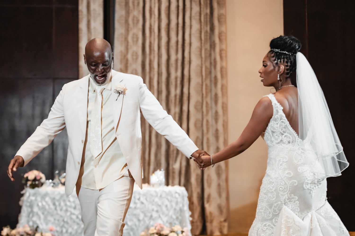 Bride in lace mermaid gown and groom in white suit share first dance at elegant wedding reception.
