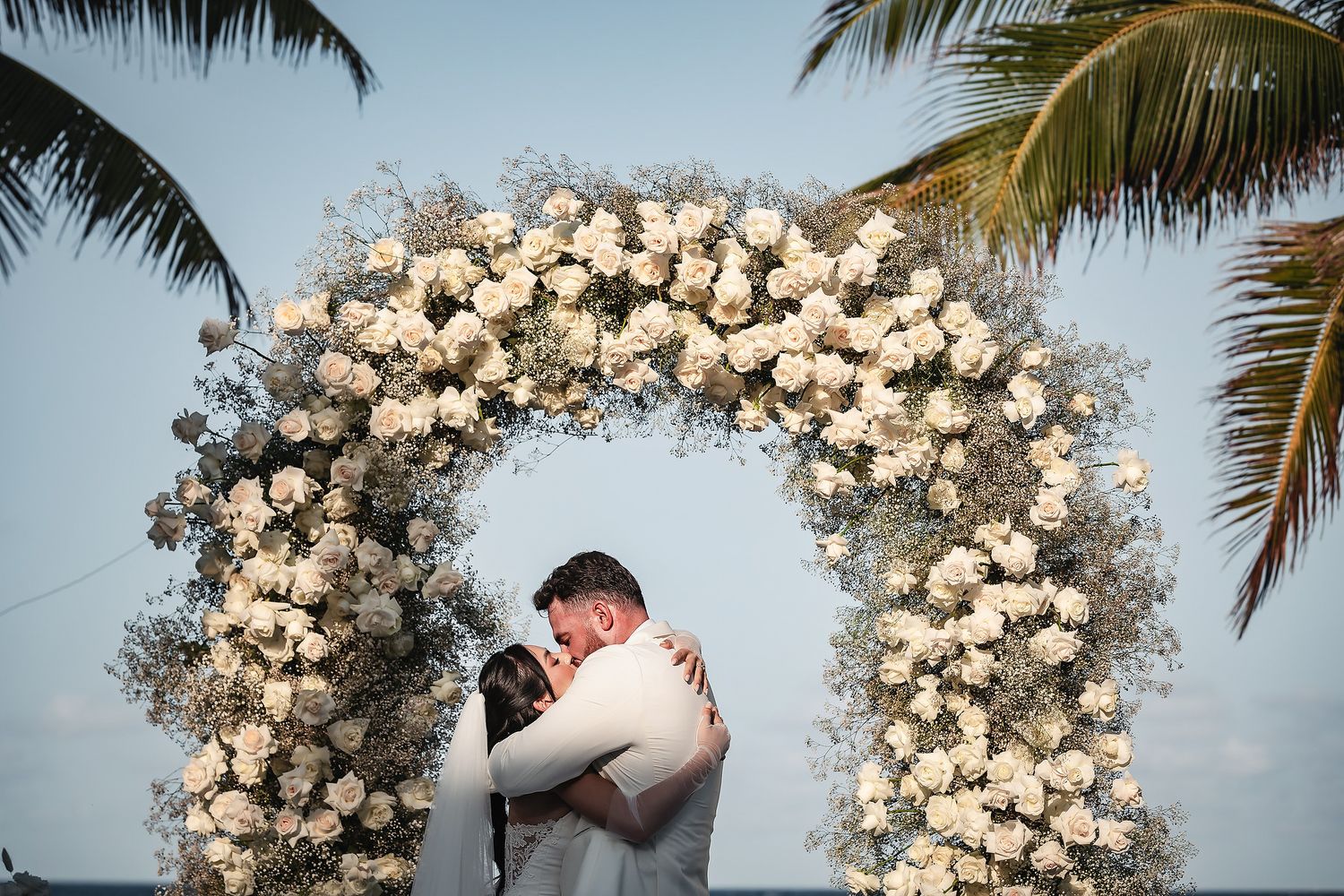 Couple embracing under a stunning white floral arch with roses and baby's breath at a beach wedding.
