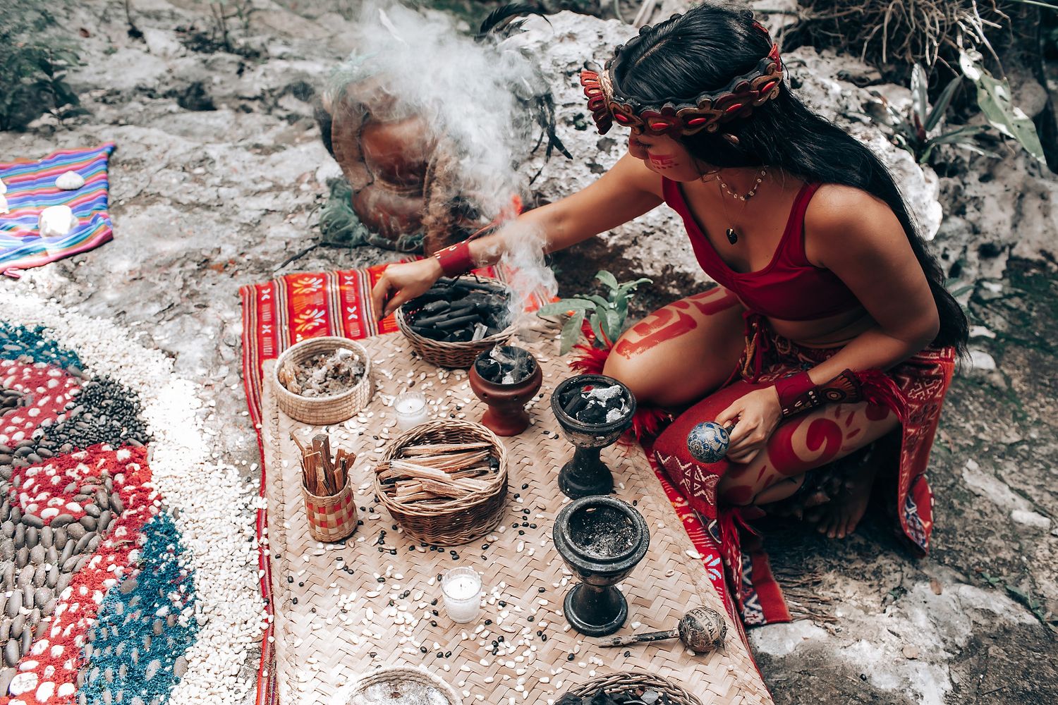Woman in red performing a traditional Mayan ritual with smoking ceremonial bowls on a colorful textile outdoors.