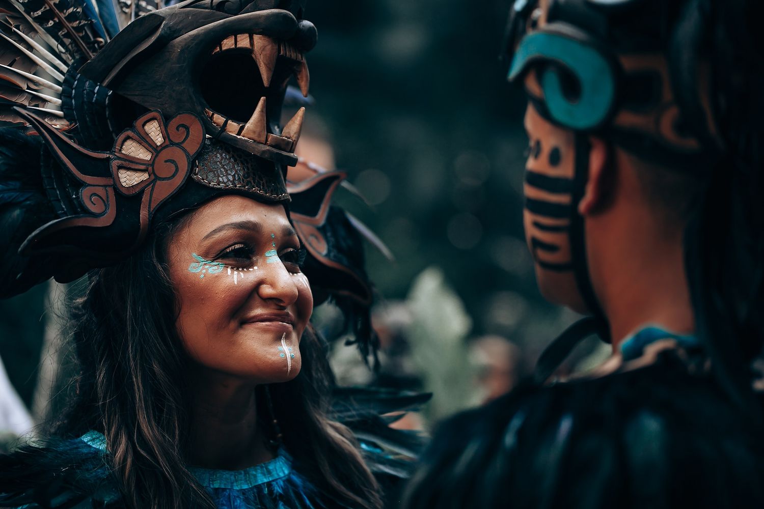Close-up of a smiling woman in Mayan ceremonial makeup and headdress facing another costumed performer.