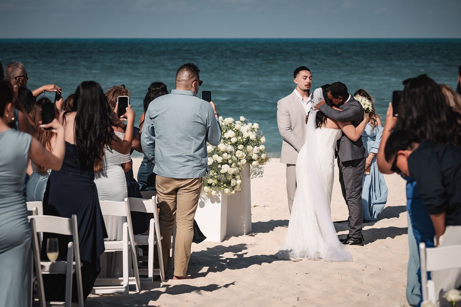 Bride and groom share first kiss at romantic beach wedding ceremony with ocean backdrop and white floral arrangements.