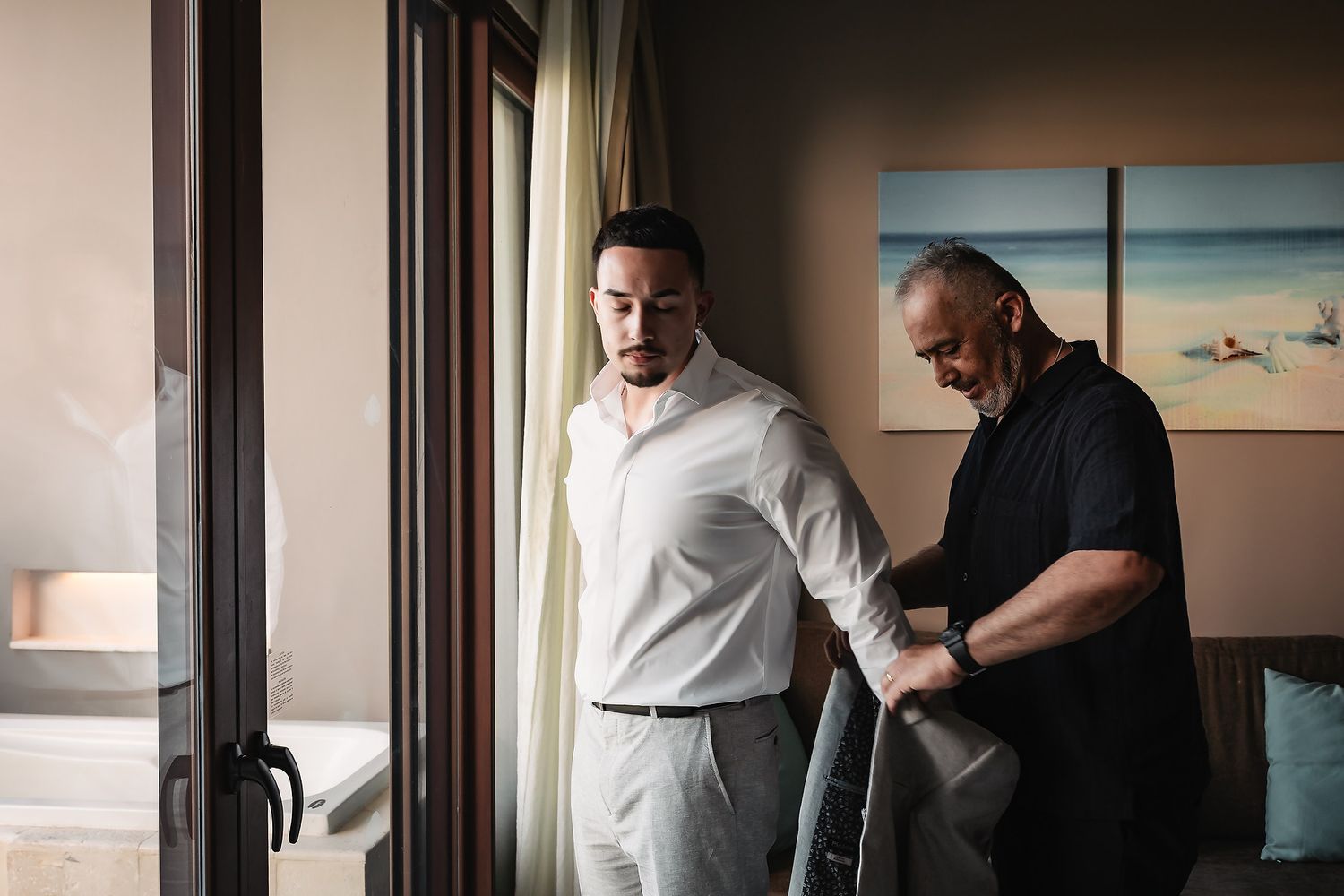 Two men in a hotel room, one in a white shirt adjusting his suit jacket near a window with curtains.