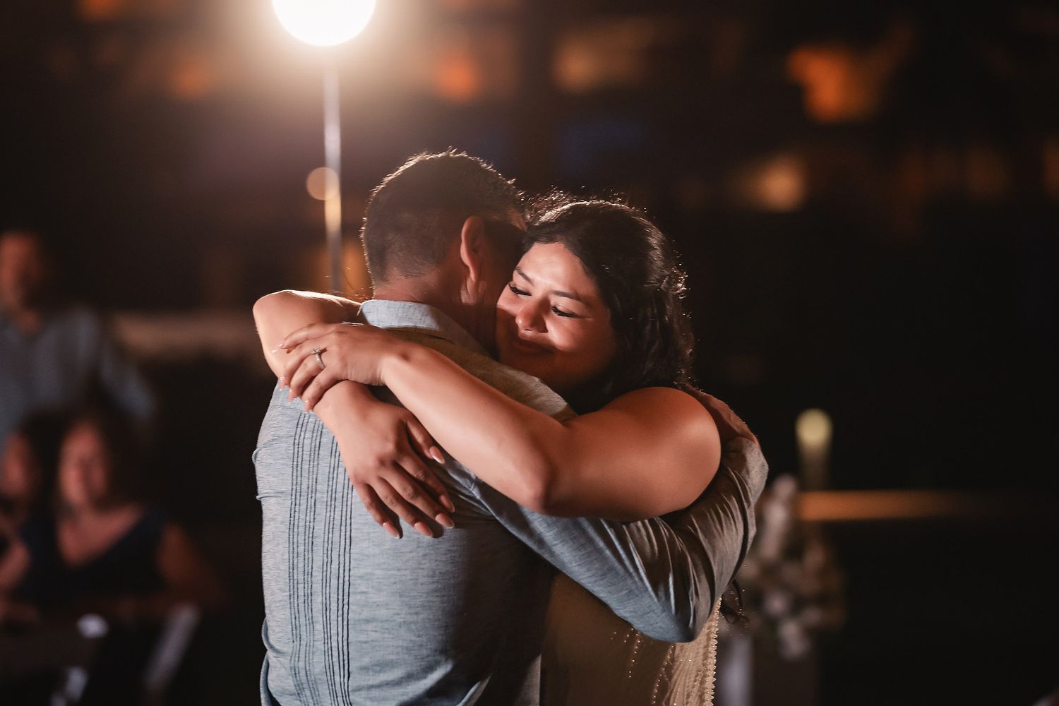 Couple sharing a heartfelt hug during their first dance at a romantic wedding reception under warm stage lighting.