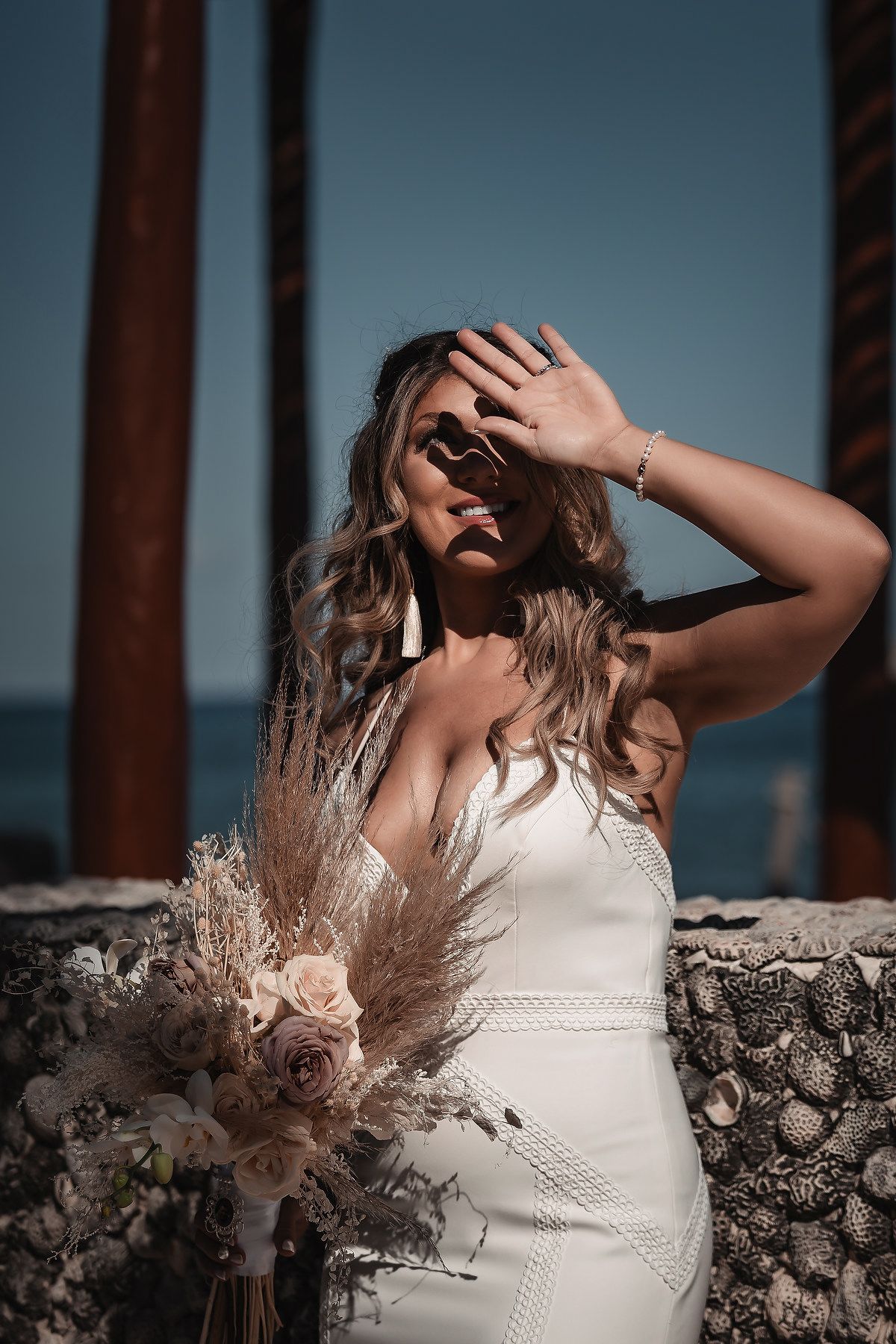 Bride in white gown shielding sun, holding boho bouquet with pampas grass and roses near ocean.