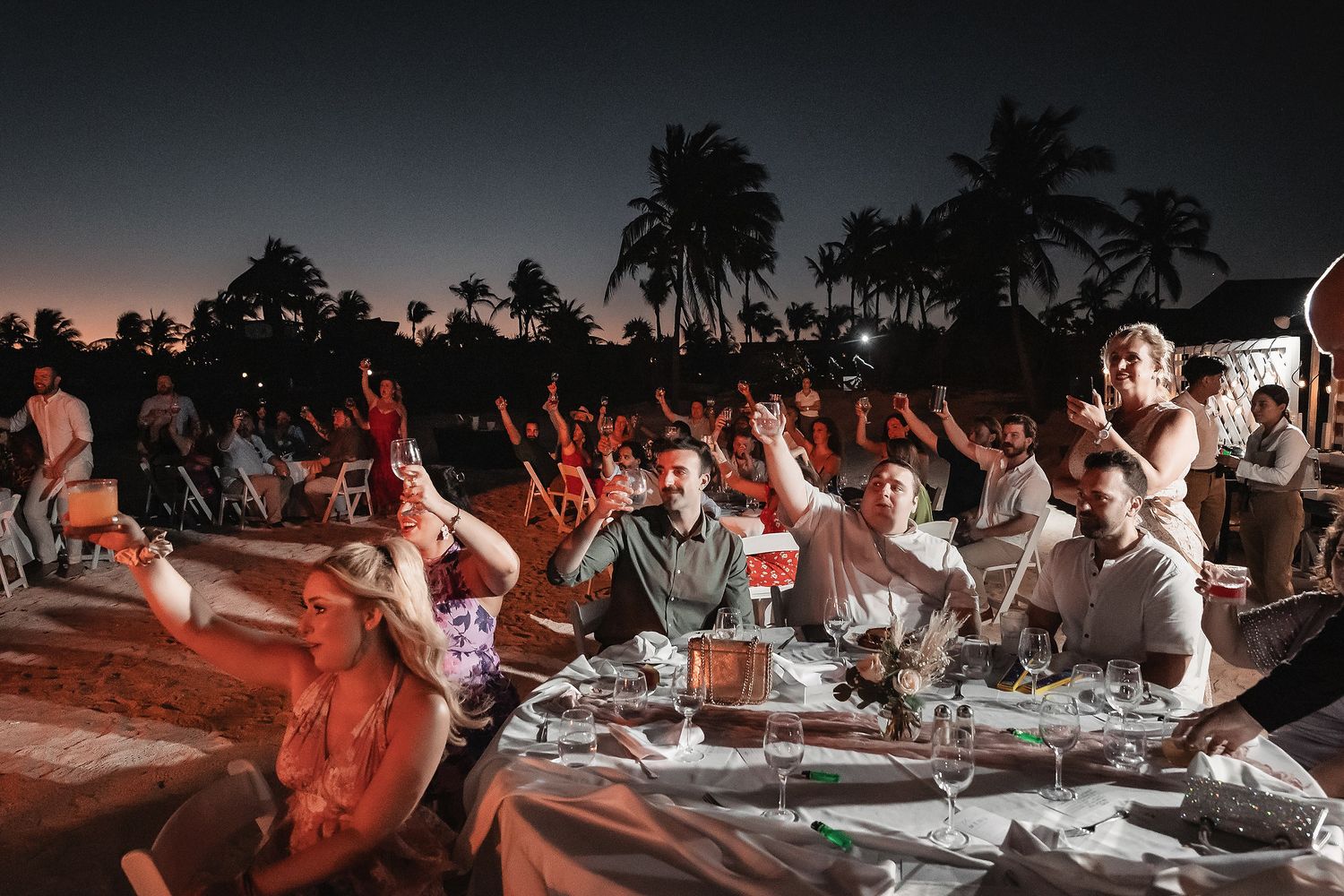 Guests celebrate at an outdoor evening dinner party under palm trees with raised hands and festive table settings.