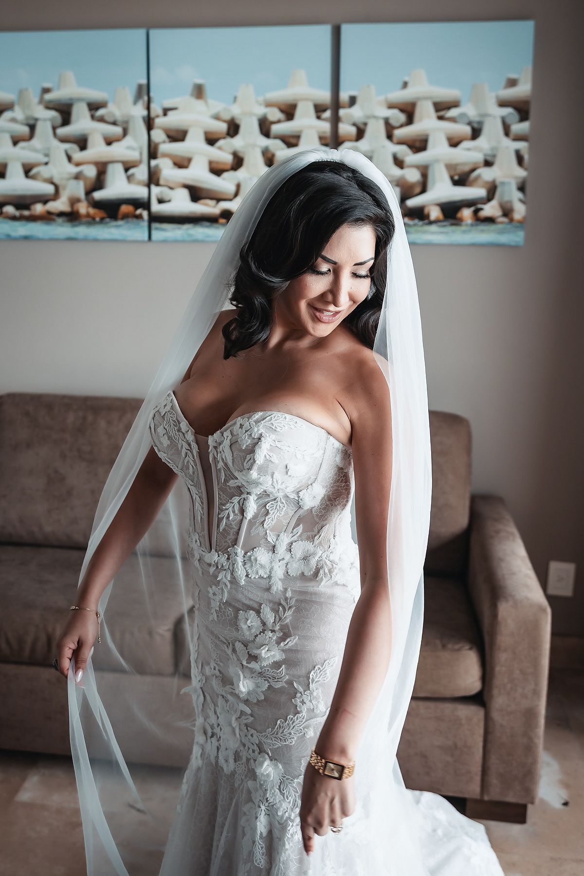 Smiling bride in strapless lace mermaid gown with flowing veil, posing indoors with coastal artwork in background.