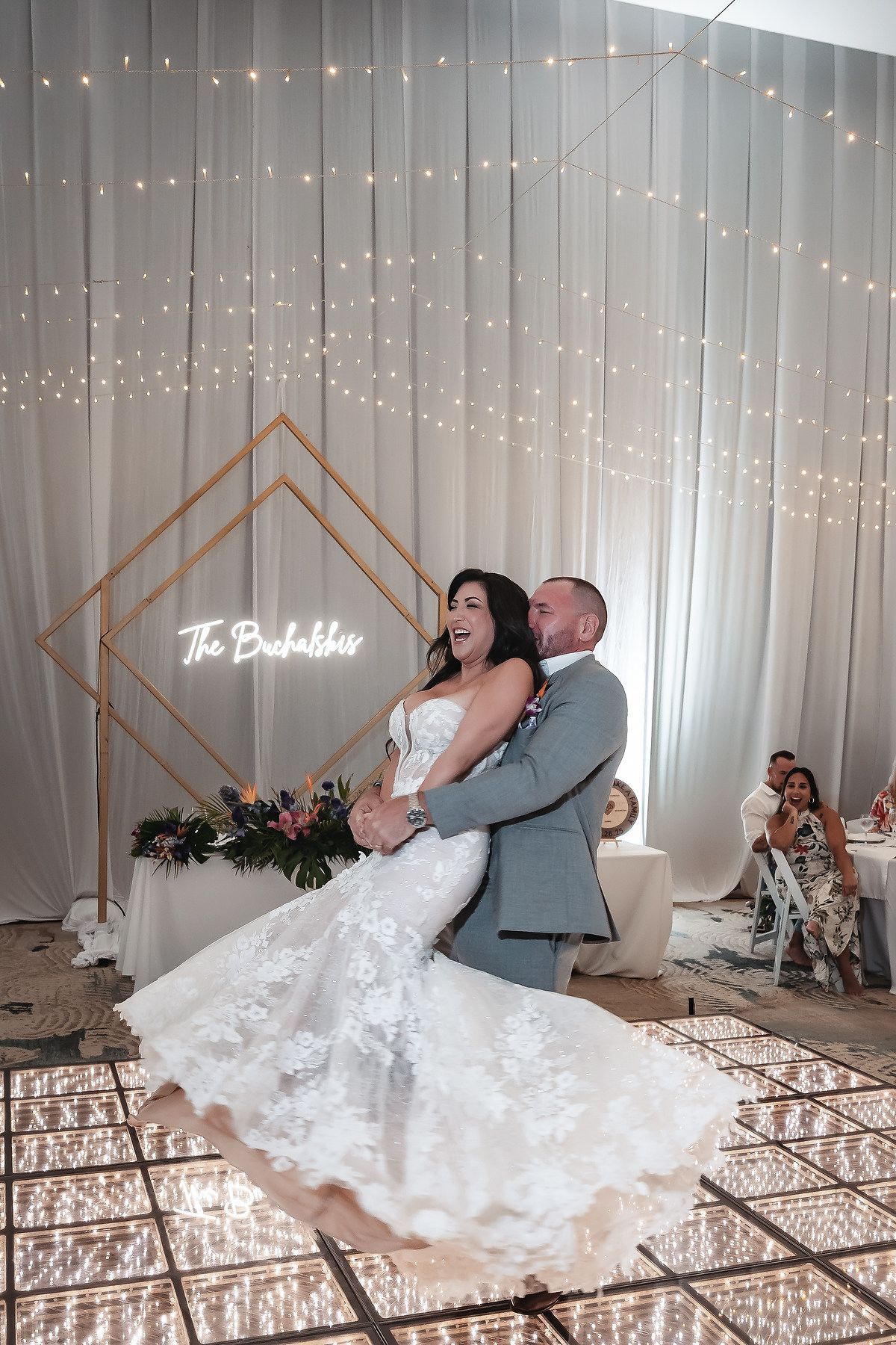 A bride and groom share a joyful first dance on a mirrored floor beneath a gold geometric arch and twinkling fairy lights.