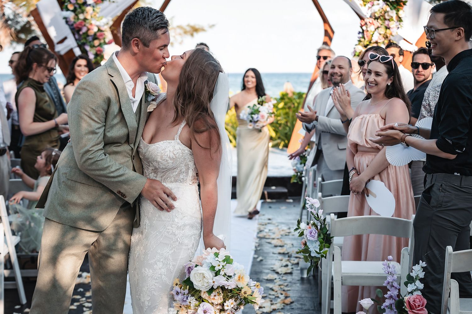 Newlyweds share first kiss at outdoor beach wedding ceremony surrounded by cheering guests.