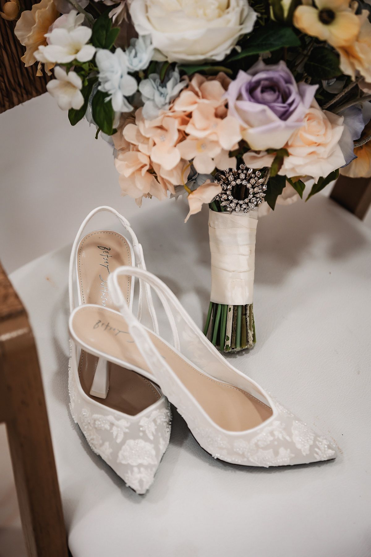 White lace bridal heels with slingback straps beside a pastel wedding bouquet with lavender roses and a brooch.