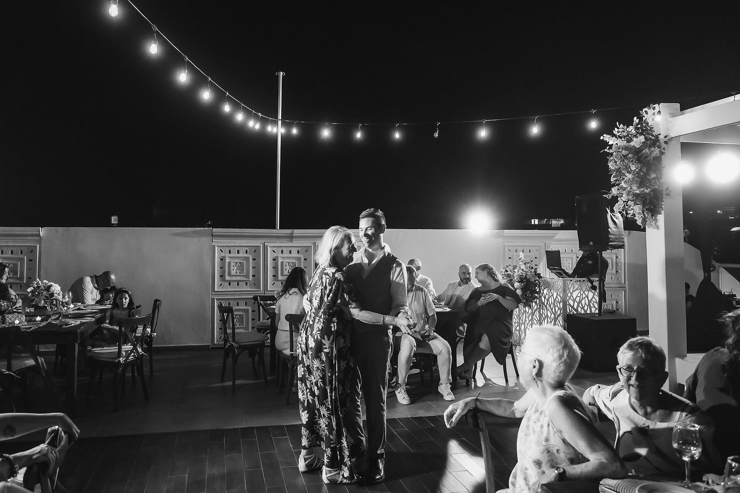 Couple sharing first dance on rooftop venue at night with string lights and guests seated nearby.