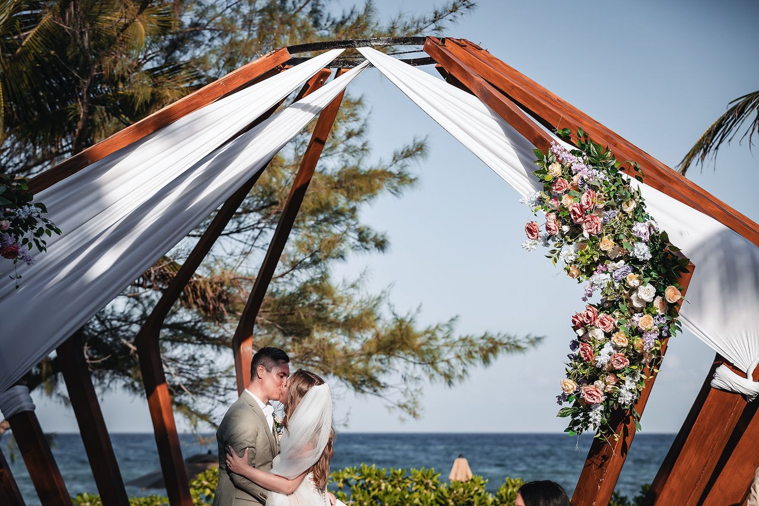 Couple kissing under a floral wooden arch at a beachside wedding ceremony with ocean views.