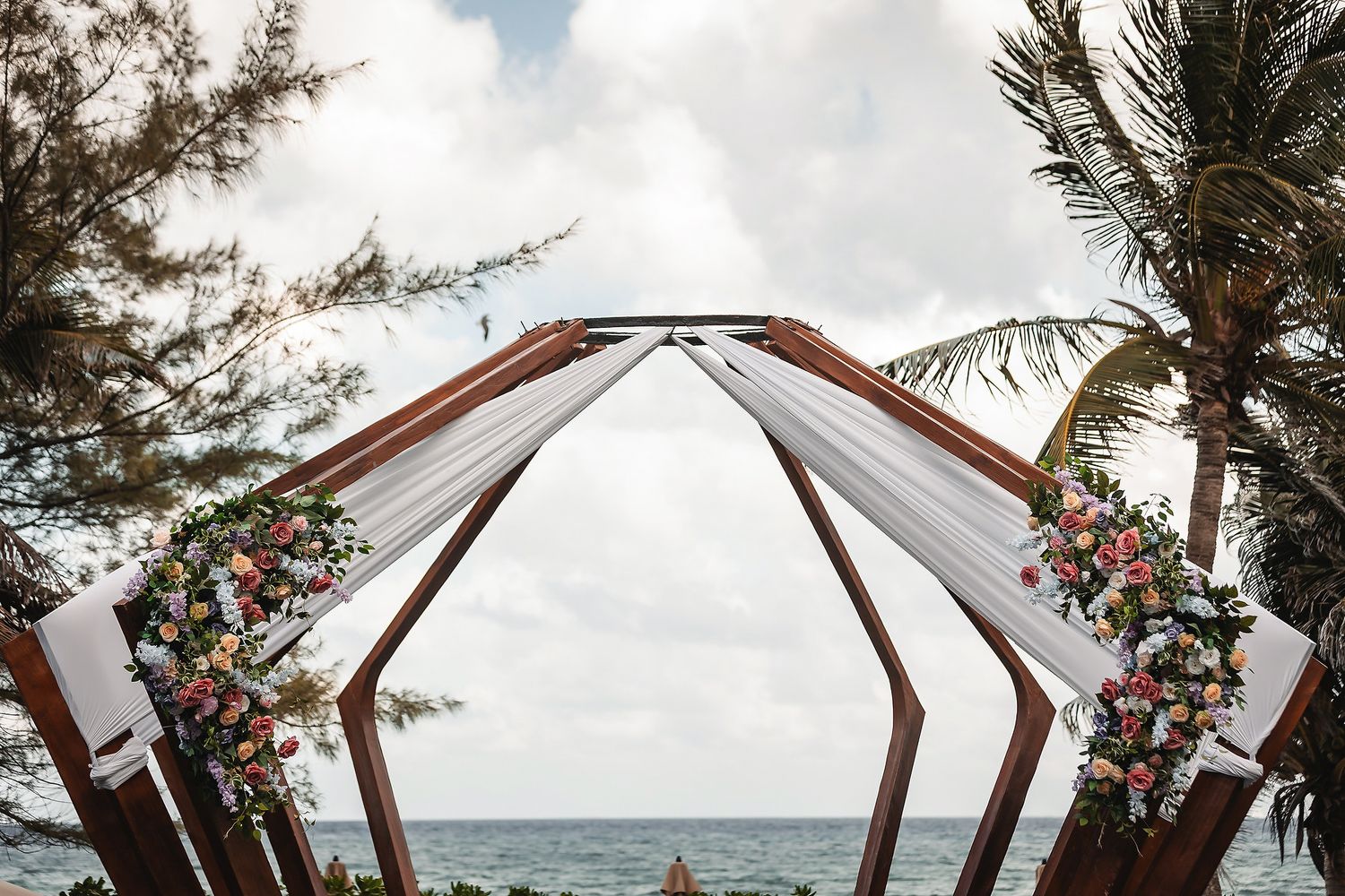 Geometric wooden wedding arch adorned with colorful floral arrangements and white draping overlooking a tropical ocean.