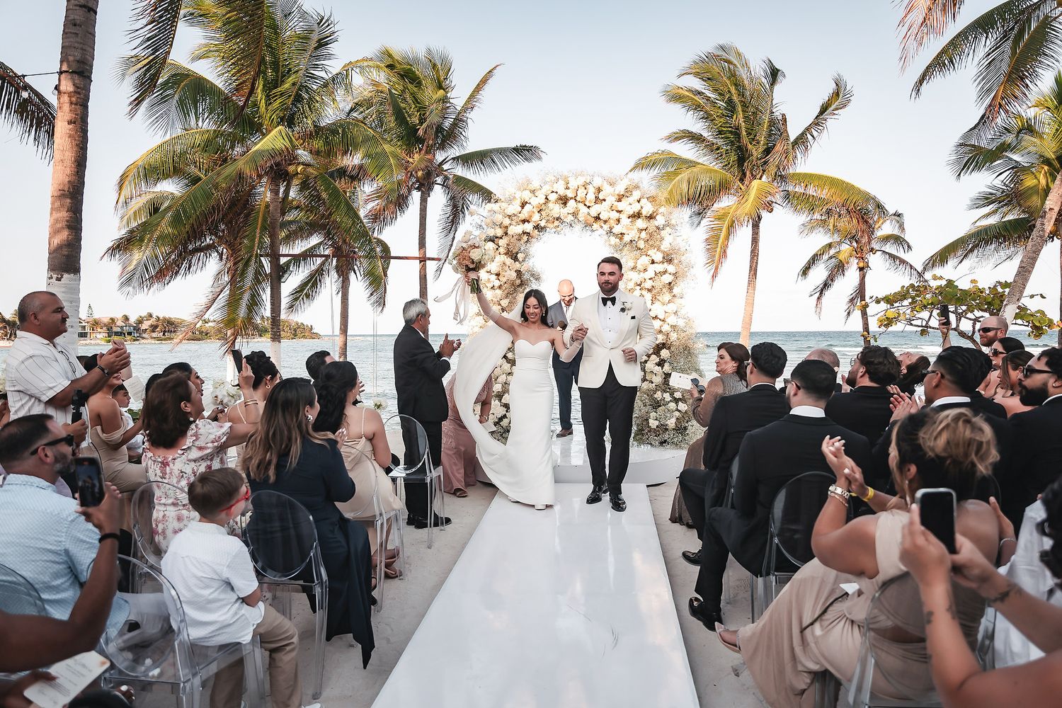 Bride and groom celebrate recessional on white aisle at tropical beach wedding with floral arch and palm trees.