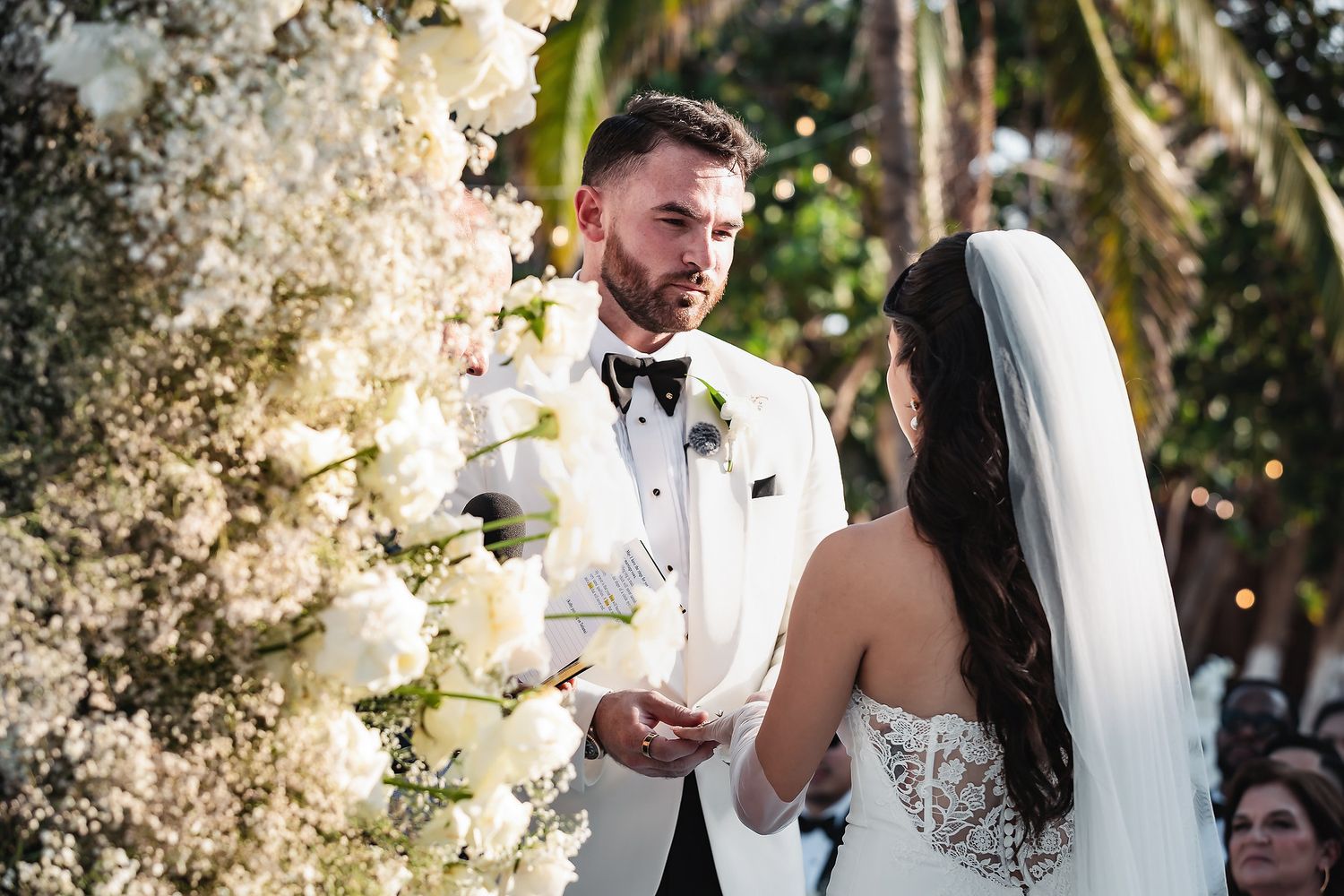 Groom in white tuxedo exchanges vows with bride in lace dress at elegant outdoor wedding ceremony with white floral arch.