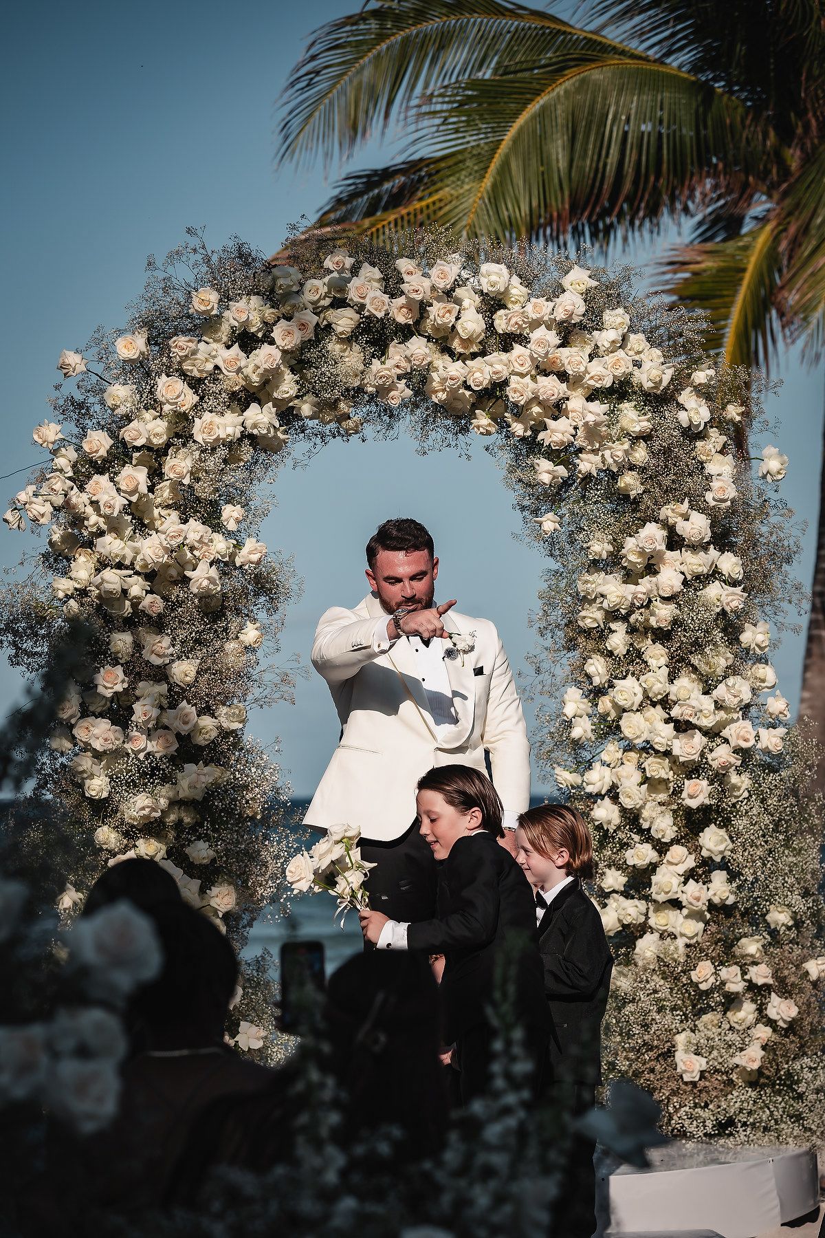Groom in white suit stands under floral arch with two boys in tuxedos at outdoor wedding ceremony.
