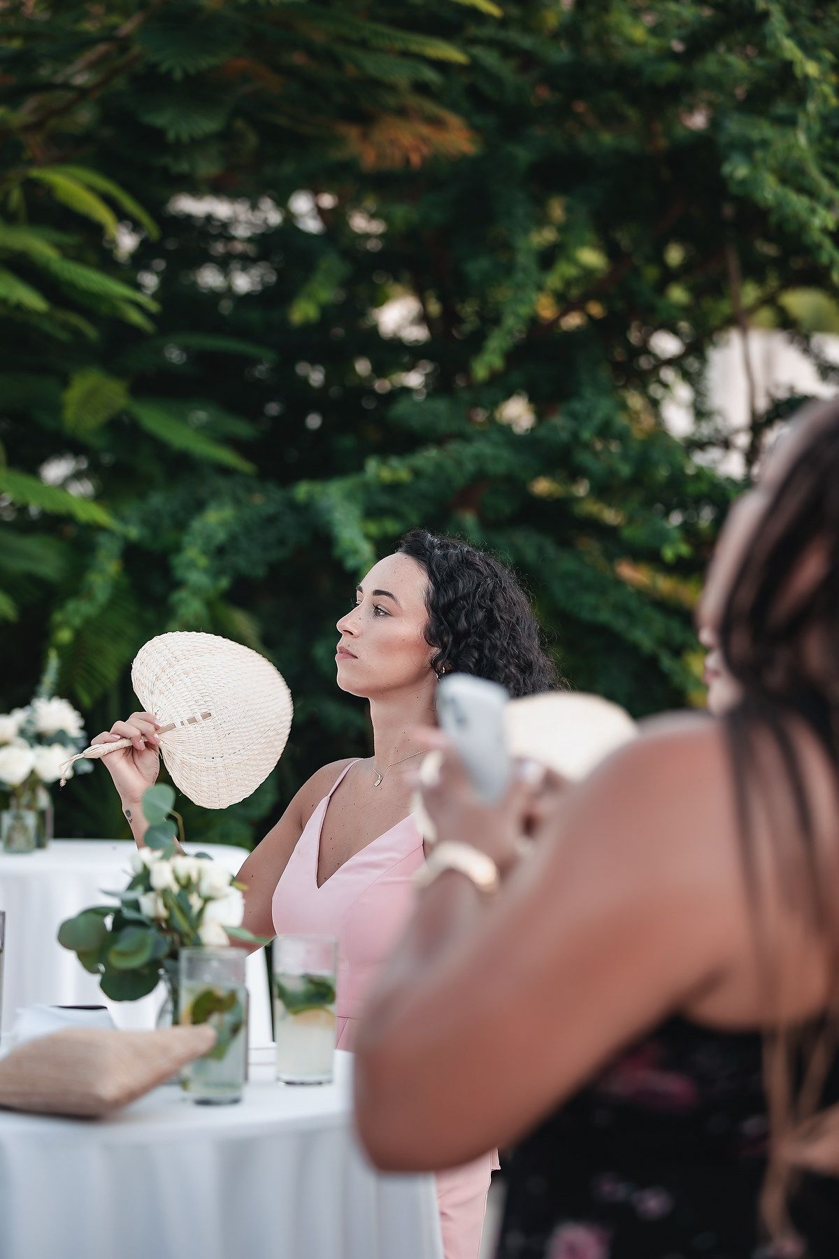 Woman in pink dress fans herself with a hand fan at an outdoor garden wedding reception on a warm day.
