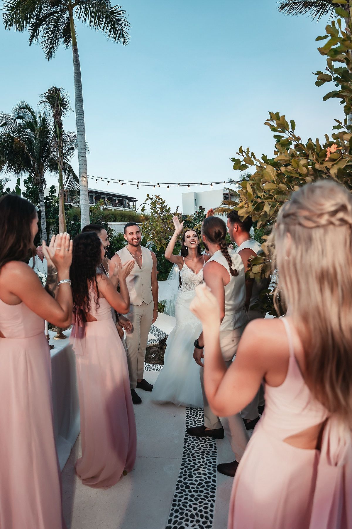Bride and groom celebrate at outdoor tropical wedding ceremony with bridesmaids in pink dresses clapping along a white aisle.