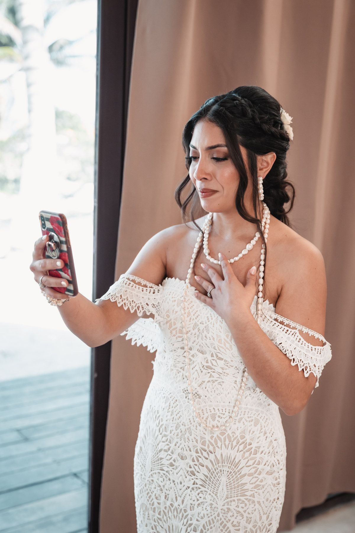 Beautiful dark-haired bride in lace off-shoulder gown taking selfie wearing pearl jewelry.