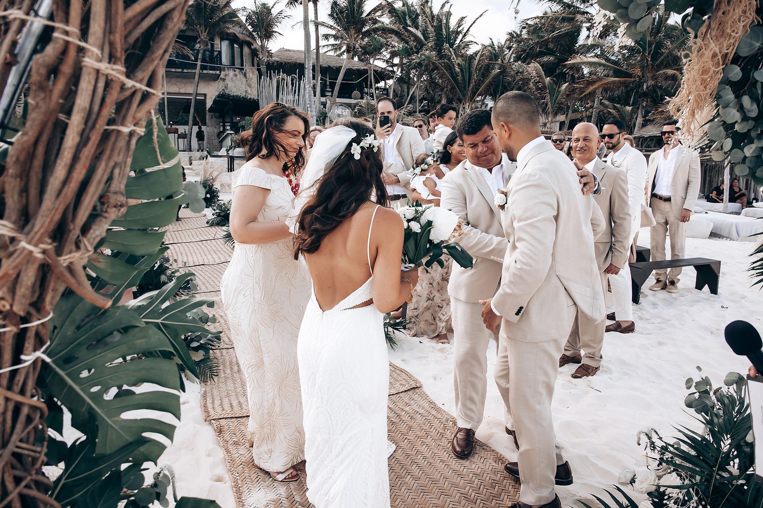 Wedding guests in white attire dance and celebrate at a tropical outdoor reception with palm trees.
