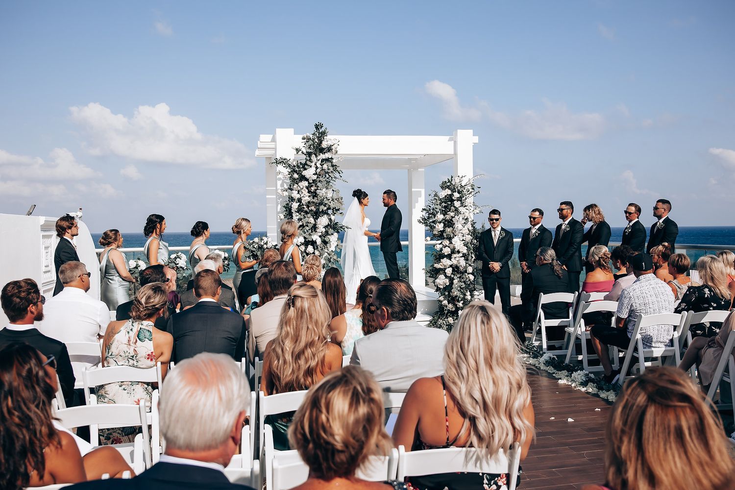Oceanfront wedding ceremony setup with white chairs and floral arch against blue sky.