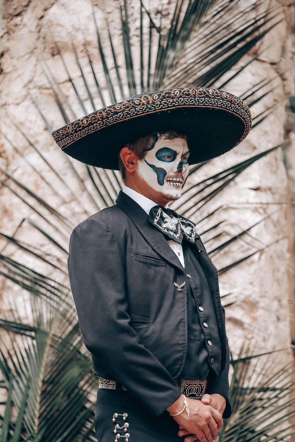 Person in black suit and wide brimmed hat with skull face paint poses against palm fronds in dramatic portrait.