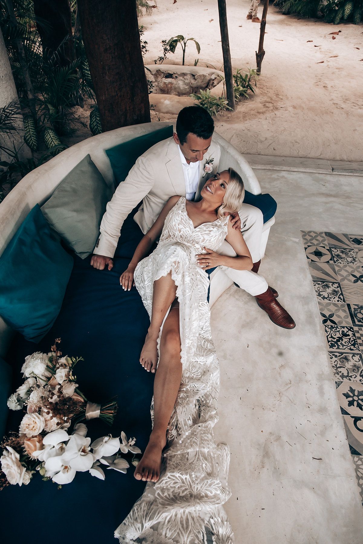 A couple in wedding attire rests together on a light-colored floor with white floral arrangements nearby.