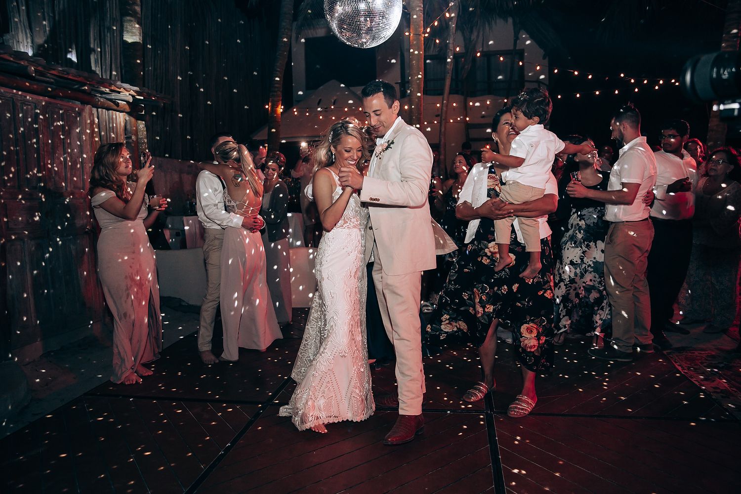 Couples dancing together on a dark dance floor illuminated by a disco ball and string lights during a wedding reception.