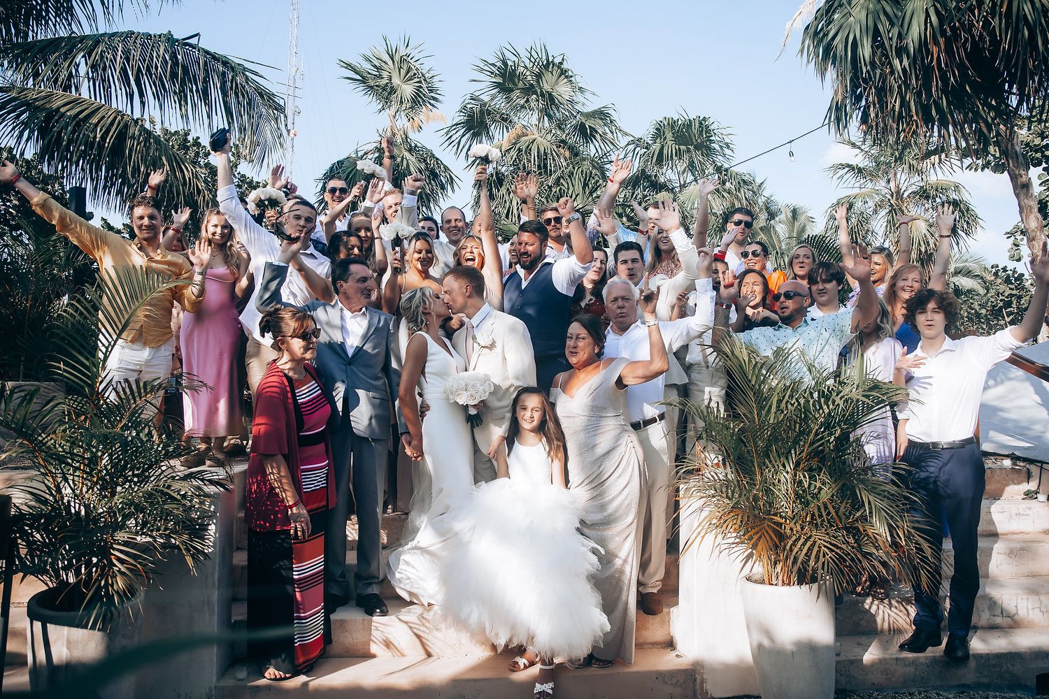 Wedding guests celebrate and cheer during a group photo among tropical palm trees.