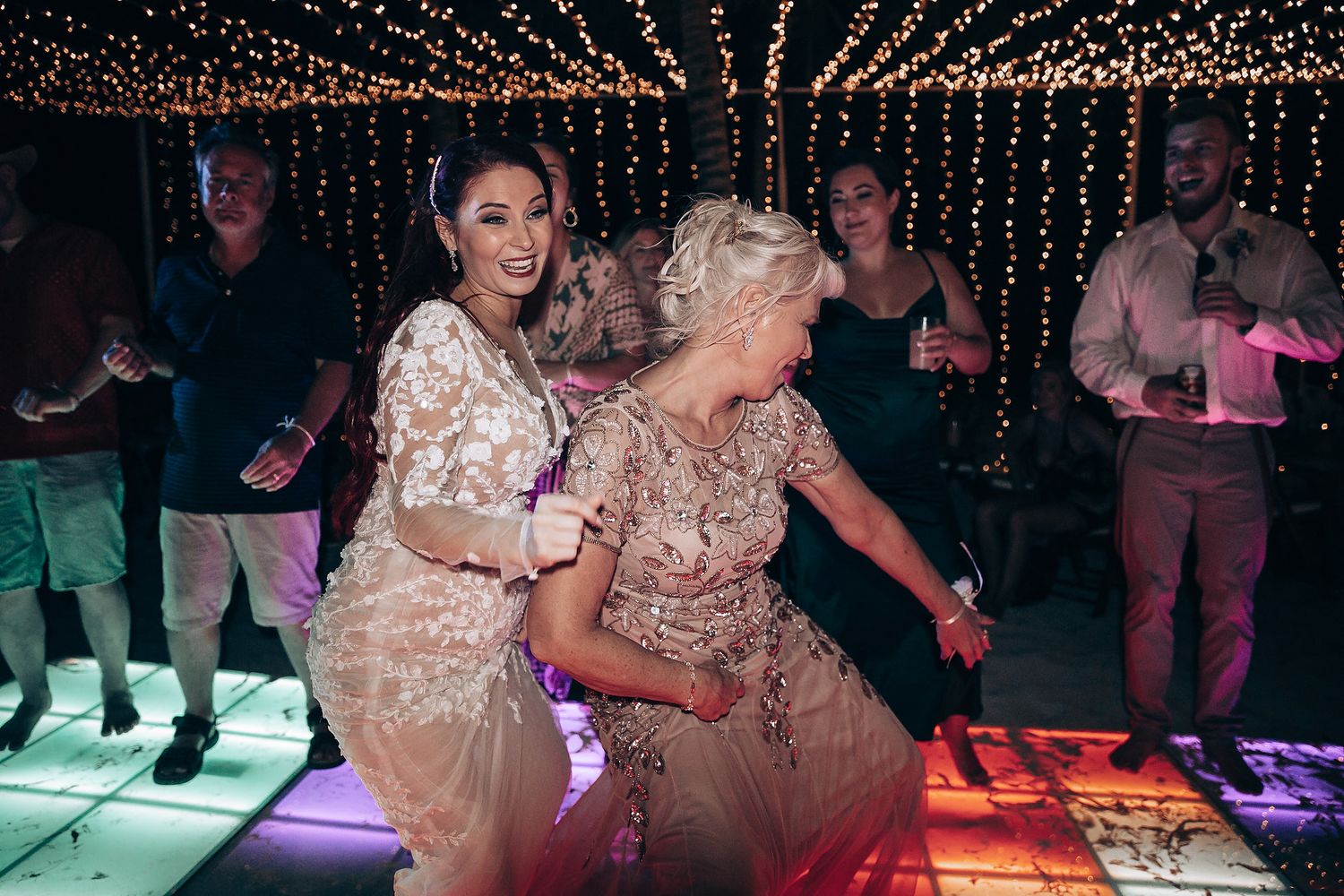Wedding reception guests dance under twinkling string lights on a colorfully lit dance floor.