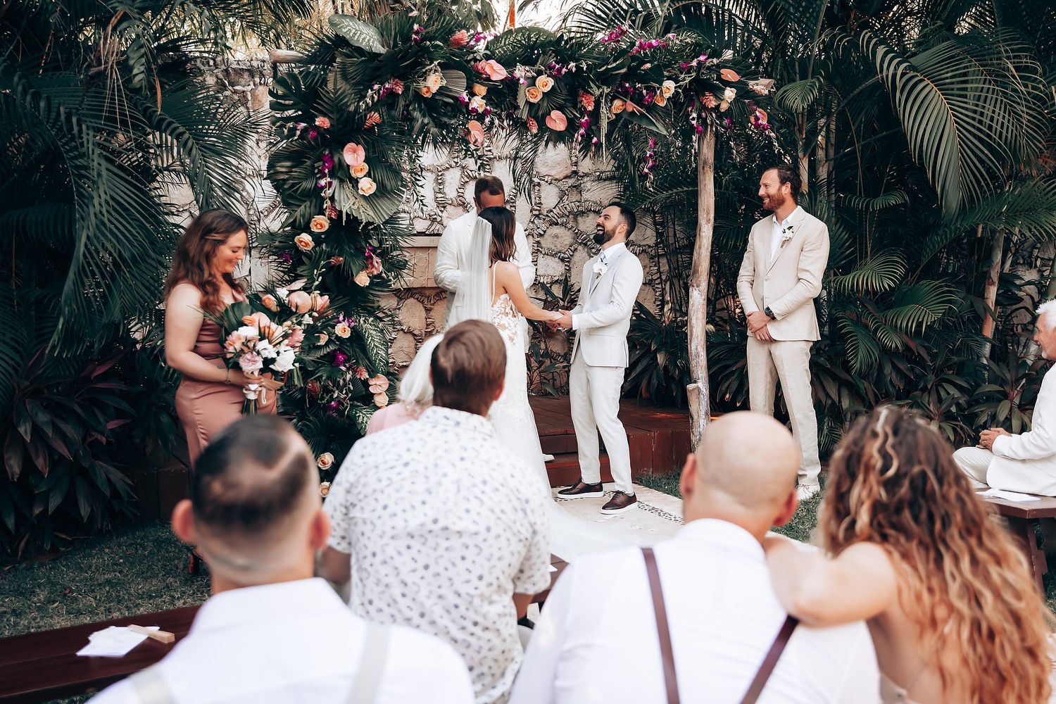 Outdoor wedding ceremony in tropical garden setting with white-clad guests and floral decorations.