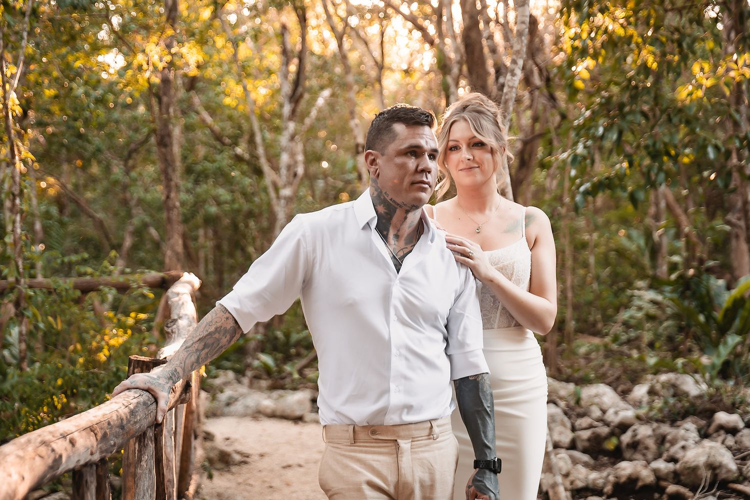 Couple in white attire standing on a wooden bridge surrounded by lush tropical forest at golden hour.