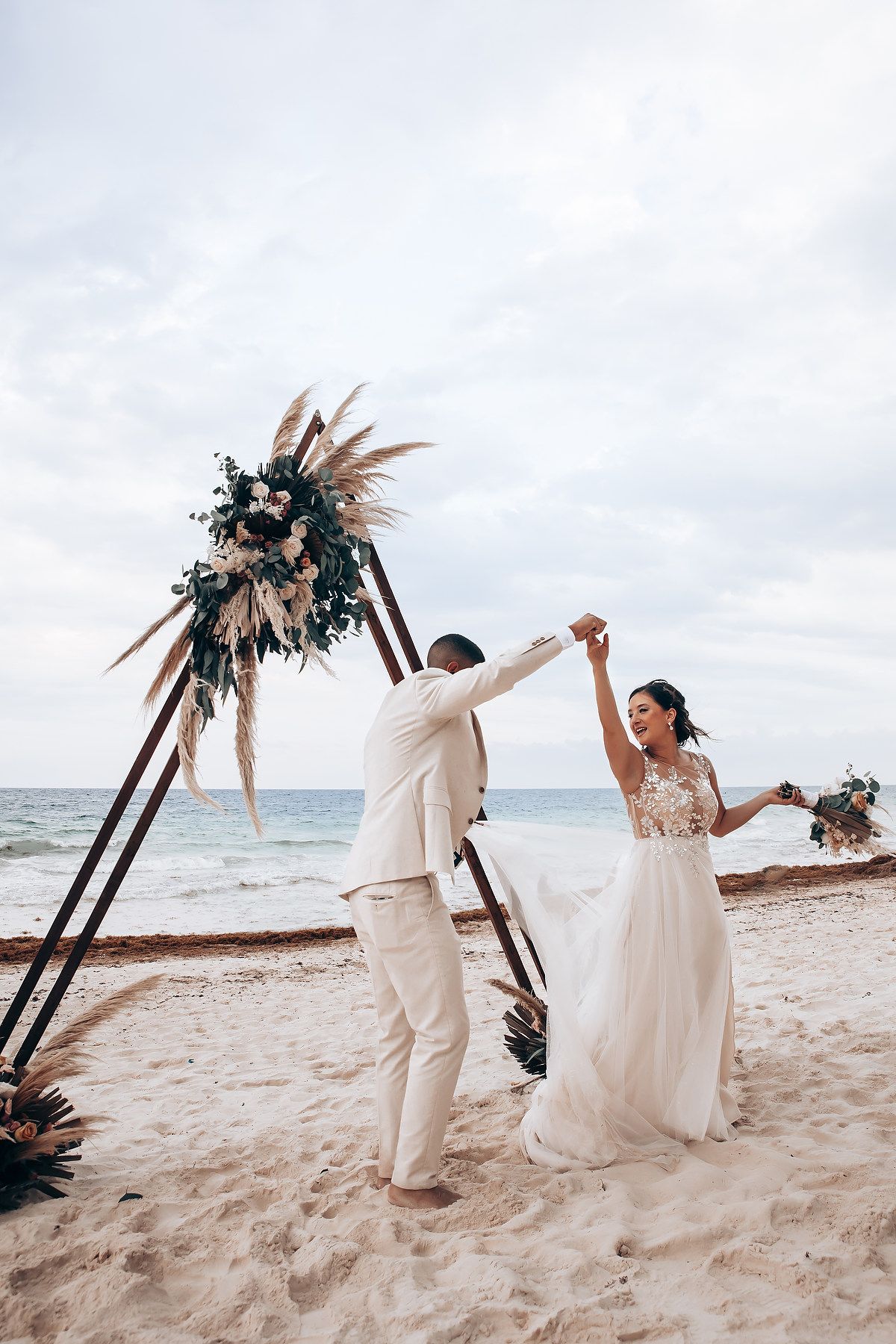 Newlyweds dancing and celebrating on beach with wooden arch and ocean backdrop.