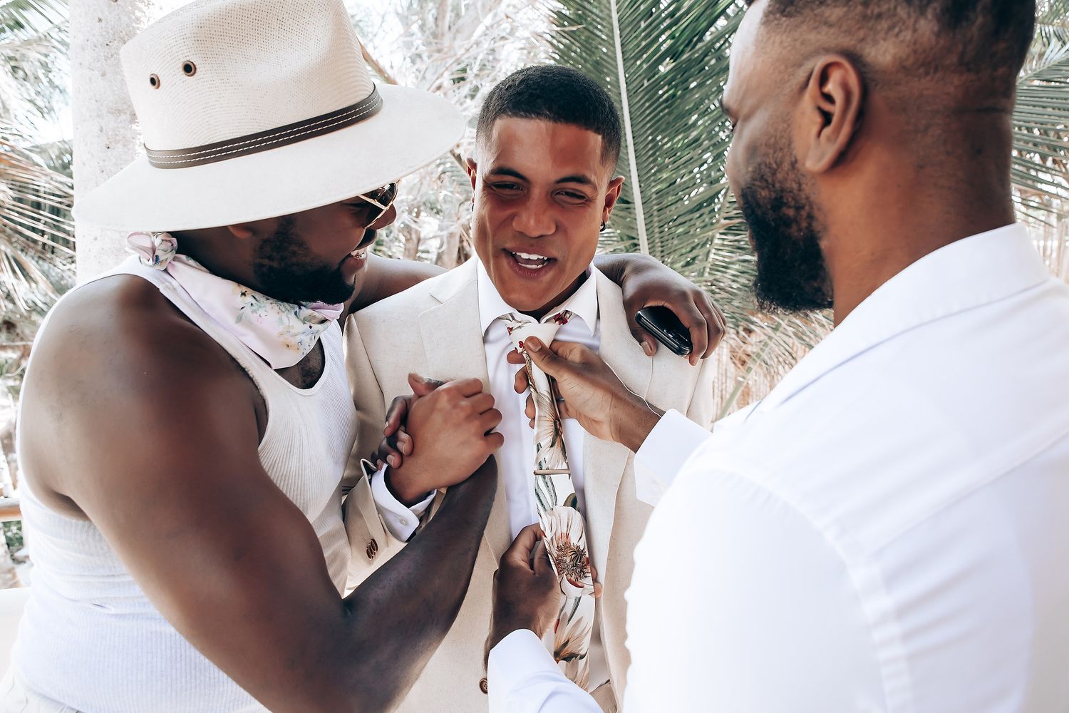 Groomsmen help adjust the groom's bowtie and attire before a tropical beach wedding ceremony.