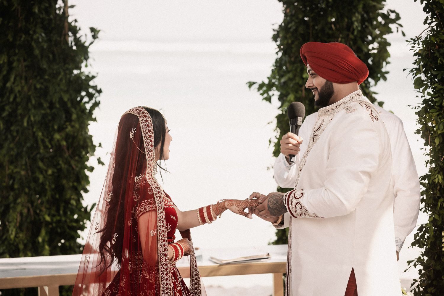 Sikh bride and groom exchange vows holding hands at a lush green outdoor wedding ceremony.