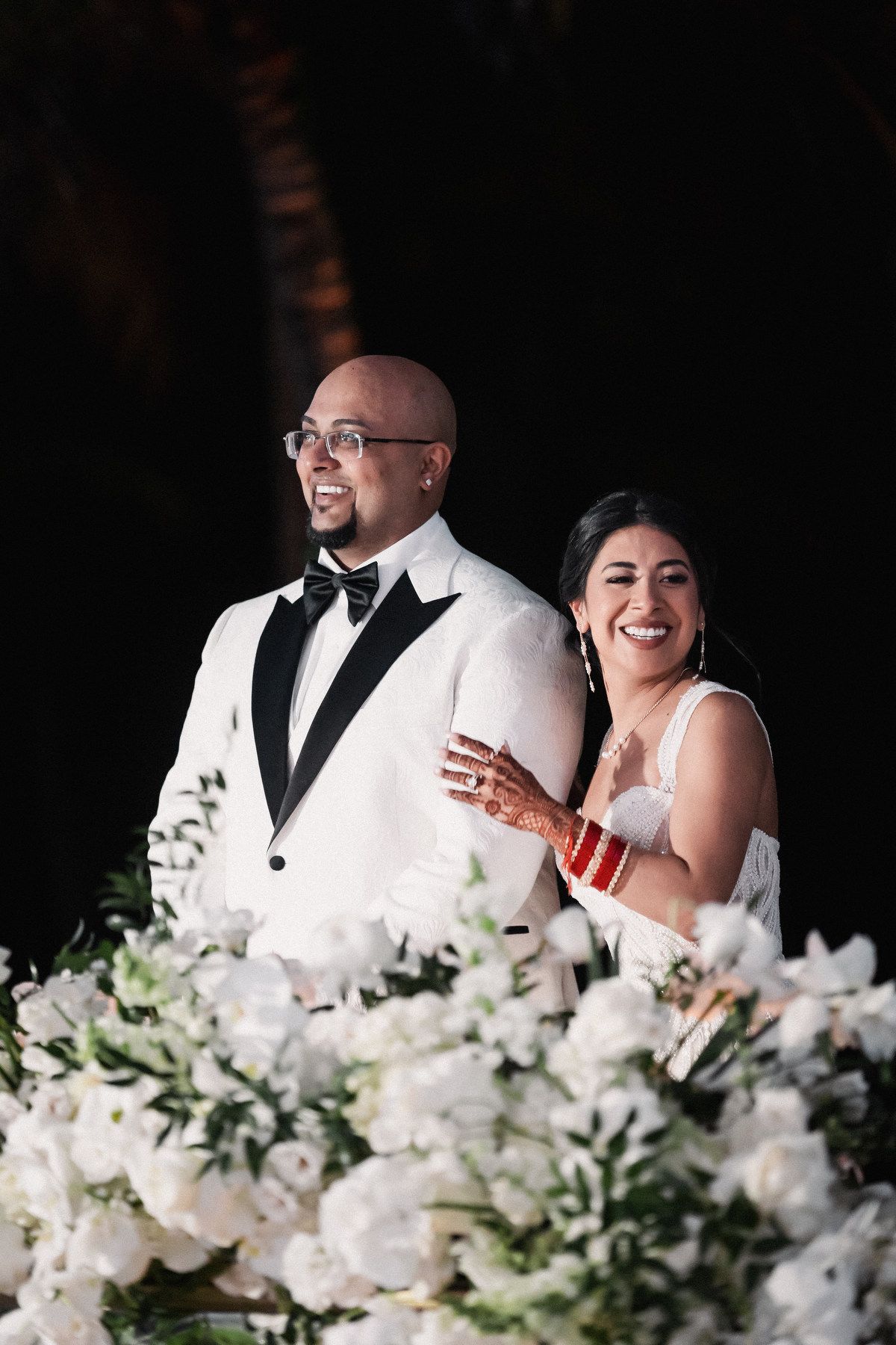 Smiling bride and groom in white wedding attire stand together surrounded by white floral arrangements at reception.