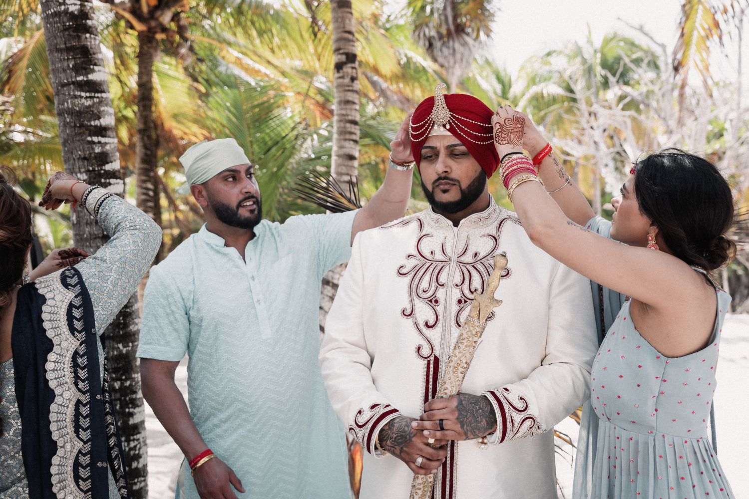 Groom in white sherwani and red turban being dressed by attendants outdoors among palm trees.
