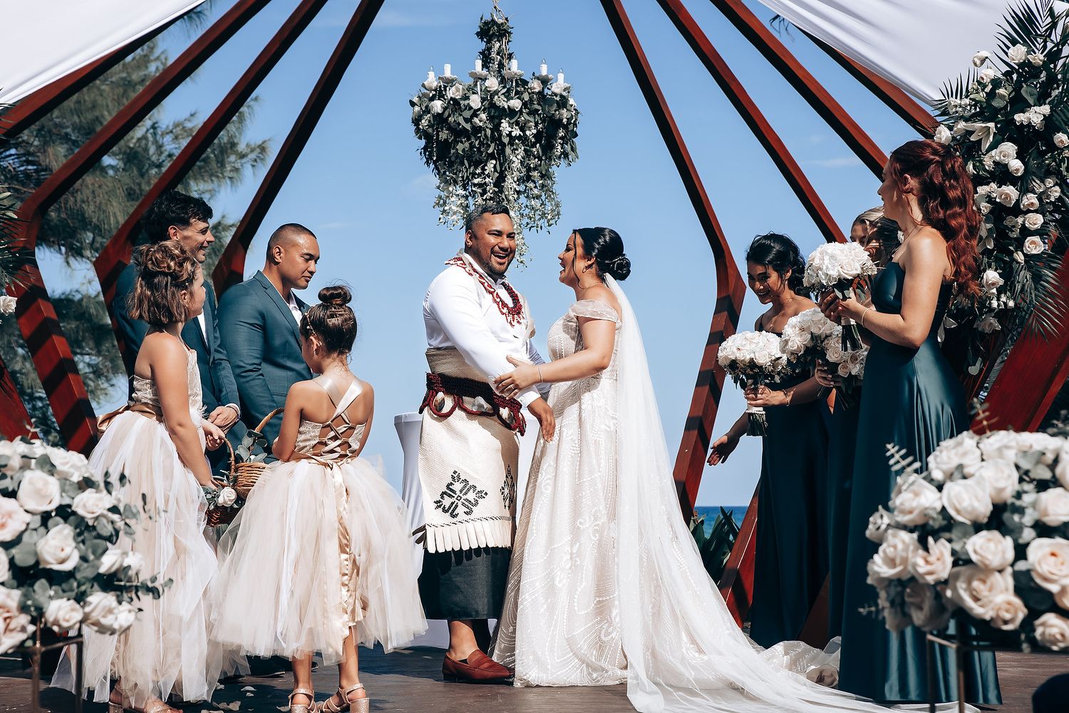 Wedding ceremony taking place under a wooden gazebo decorated with white flowers and greenery.