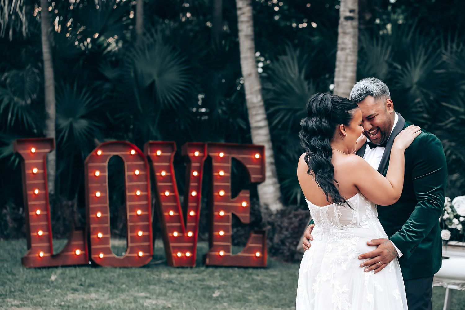 Elegant couple embraces during outdoor wedding photoshoot with illuminated LOVE sign in background.
