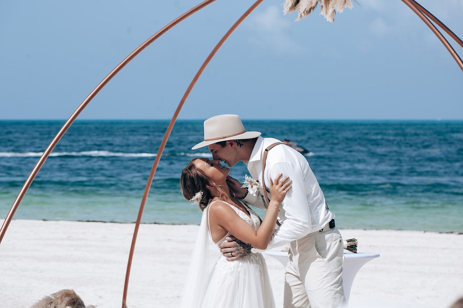 A romantic moment during a beach wedding ceremony under a copper arch with ocean backdrop.