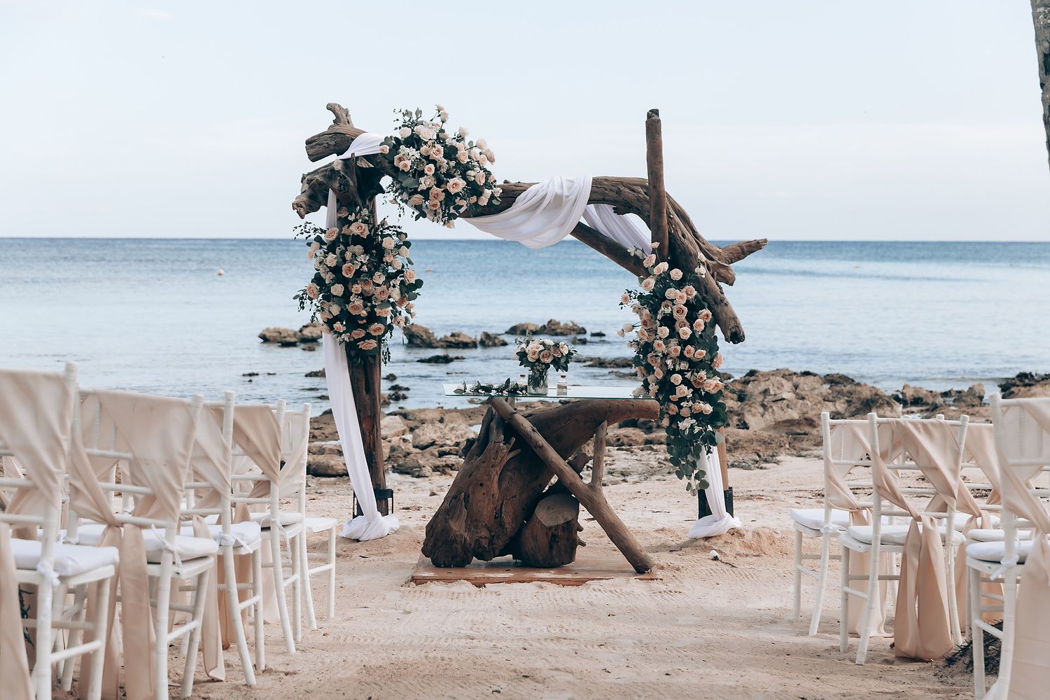 White chairs set up for a beach wedding ceremony with ocean views and tropical decor.